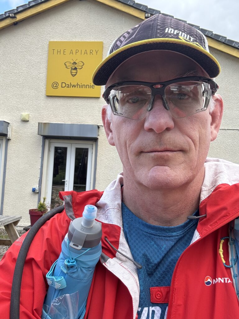 Picture of me in front of The Apiary, wearing my Infinit cap, an orange rain jacket, and hydration vest with chest bottles
