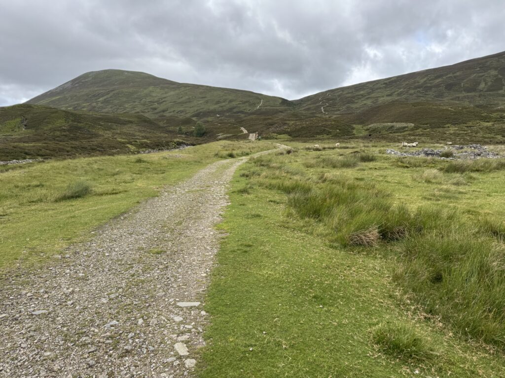 A gravel path in the foreground, with steep green hills in the background. 