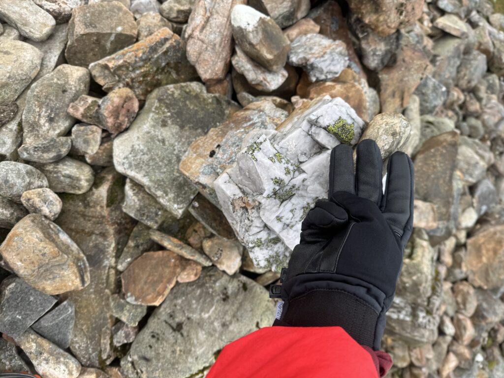 Tapping the summit rock pile with a black glove, showing three fingers.